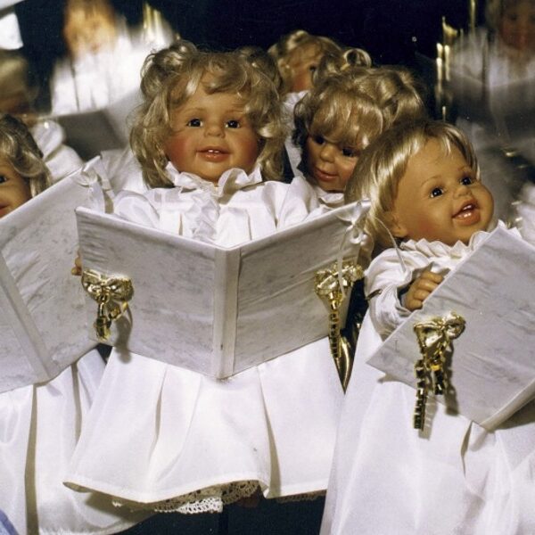 Vier Puppen mit blondem Haar, gekleidet in weiße Gewänder, halten Liederbücher in der Hand, als ob sie in einem Chor singen würden.