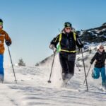 Drei Personen, die auf einer verschneiten Piste Skilanglauf betreiben, tragen Winterkleidung und Sonnenbrillen, mit einem Gebäude und einem Berg im Hintergrund.