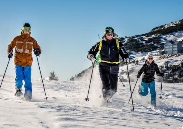 Drei Personen, die auf einer verschneiten Piste Skilanglauf betreiben, tragen Winterkleidung und Sonnenbrillen, mit einem Gebäude und einem Berg im Hintergrund.