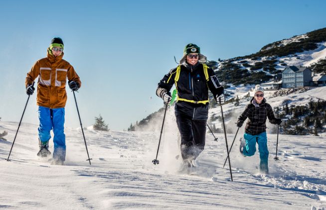 Drei Personen, die auf einer verschneiten Piste Skilanglauf betreiben, tragen Winterkleidung und Sonnenbrillen, mit einem Gebäude und einem Berg im Hintergrund.
