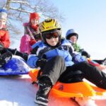 Vier in Winterkleidung gekleidete Kinder fahren an einem sonnigen Tag mit dem Schlitten einen verschneiten Hügel hinunter.