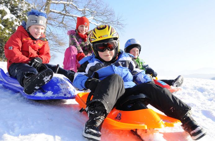 Vier in Winterkleidung gekleidete Kinder fahren an einem sonnigen Tag mit dem Schlitten einen verschneiten Hügel hinunter.