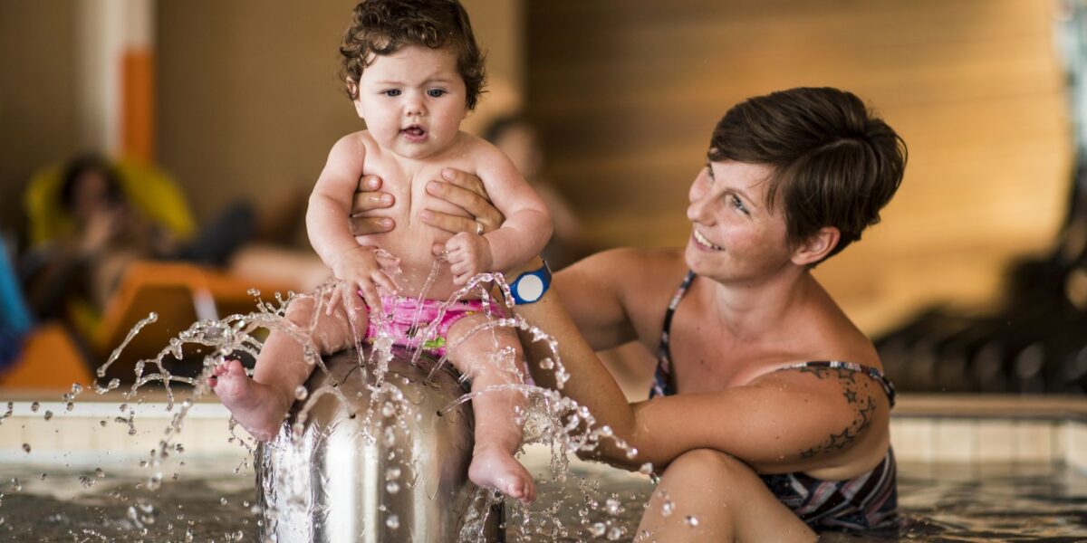 Ein Erwachsener hält ein Baby, das auf einem Springbrunnen in einem Schwimmbecken sitzt, wobei das Wasser plätschert und beide die Aktivität zu genießen scheinen.