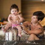 Ein Erwachsener hält ein Baby, das auf einem Springbrunnen in einem Schwimmbecken sitzt, wobei das Wasser plätschert und beide die Aktivität zu genießen scheinen.