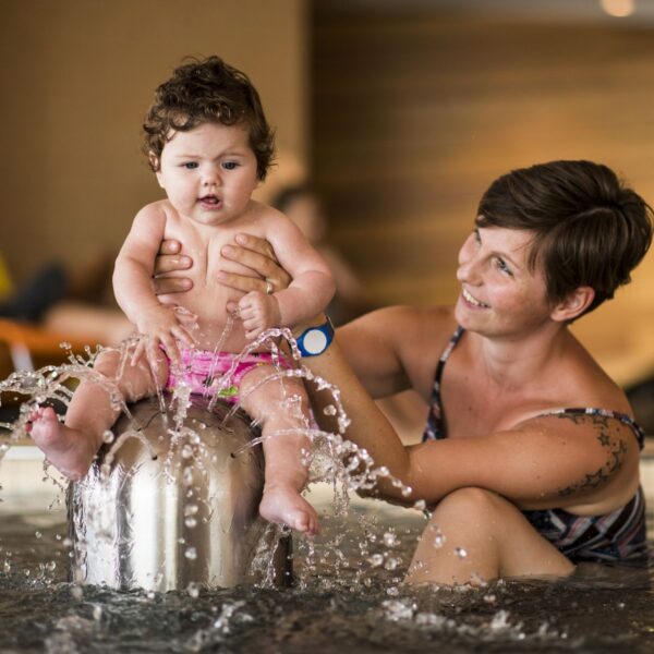 Ein Erwachsener hält ein Baby, das auf einem Springbrunnen in einem Schwimmbecken sitzt, wobei das Wasser plätschert und beide die Aktivität zu genießen scheinen.