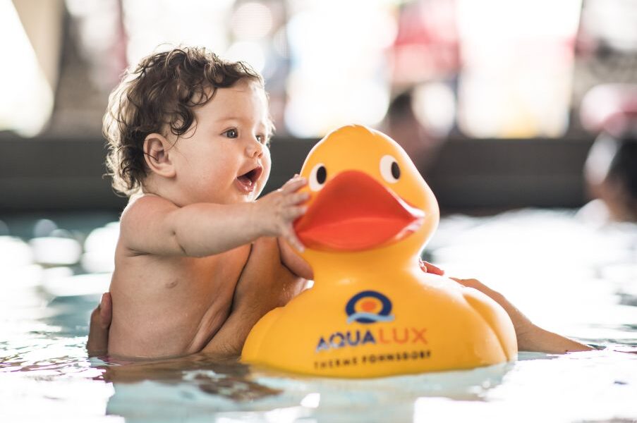 Ein Baby in einem Schwimmbecken greift nach einer großen gelben Gummiente mit der Aufschrift "AQUALUX", während es von einer Erwachsenenhand gestützt wird.