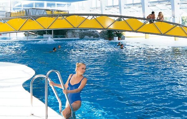 Eine Frau in einem blauen Badeanzug klettert aus einem Hallenbad in der Nähe einer gelben Brücke, während im Hintergrund mehrere Personen zu sehen sind.