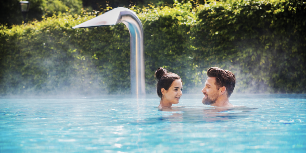 Ein Mann und eine Frau stehen sich in einem Außenpool gegenüber, aus dem Dampf aufsteigt, umgeben von Grünpflanzen und einem Wasserspiel aus Metall im Hintergrund.