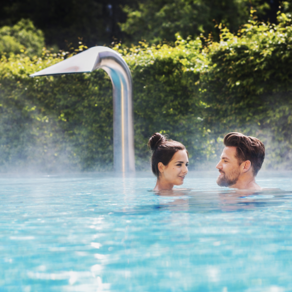 Ein Mann und eine Frau stehen sich in einem Außenpool gegenüber, aus dem Dampf aufsteigt, umgeben von Grünpflanzen und einem Wasserspiel aus Metall im Hintergrund.