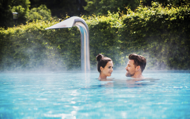 Ein Mann und eine Frau stehen sich in einem Außenpool gegenüber, aus dem Dampf aufsteigt, umgeben von Grünpflanzen und einem Wasserspiel aus Metall im Hintergrund.