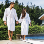 Ein Mann und eine Frau in weißen Bademänteln spazieren Hand in Hand an einem Außenpool mit Bäumen und einem modernen Gebäude im Hintergrund.