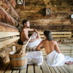 Zwei in Handtücher gehüllte Personen sitzen auf Holzbänken in einer Blockhaussauna. Im Vordergrund ist ein Holzeimer mit einer Schöpfkelle zu sehen.