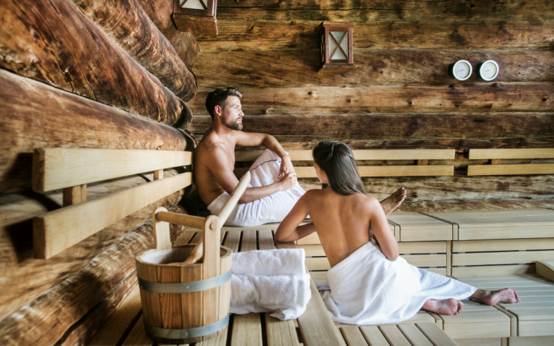 Zwei in Handtücher gehüllte Personen sitzen auf Holzbänken in einer Blockhaussauna. Im Vordergrund ist ein Holzeimer mit einer Schöpfkelle zu sehen.