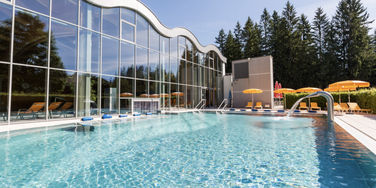 Außenschwimmbad mit klarem Wasser, gelben Liegestühlen und Sonnenschirmen neben einem modernen Glasgebäude, umgeben von Bäumen und unter freiem Himmel.