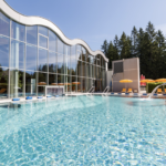 Außenschwimmbad mit klarem Wasser, gelben Liegestühlen und Sonnenschirmen neben einem modernen Glasgebäude, umgeben von Bäumen und unter freiem Himmel.