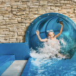Ein Kind mit einer Schwimmbrille taucht aus einer blauen Wasserrutsche auf und spritzt mit erhobenen Armen in ein Becken vor einem Steinmauerhintergrund.