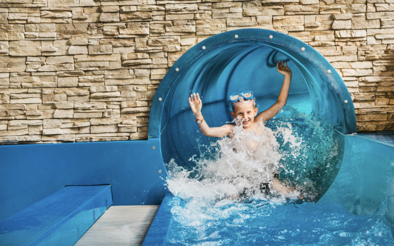 Ein Kind mit einer Schwimmbrille taucht aus einer blauen Wasserrutsche auf und spritzt mit erhobenen Armen in ein Becken vor einem Steinmauerhintergrund.