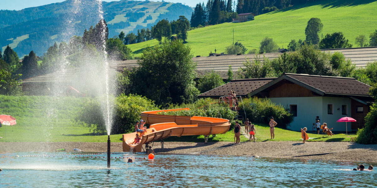 Menschen genießen einen sonnigen Tag am Strand eines Sees mit einer Wasserrutsche, einem Springbrunnen und grasbewachsenen Hügeln im Hintergrund.