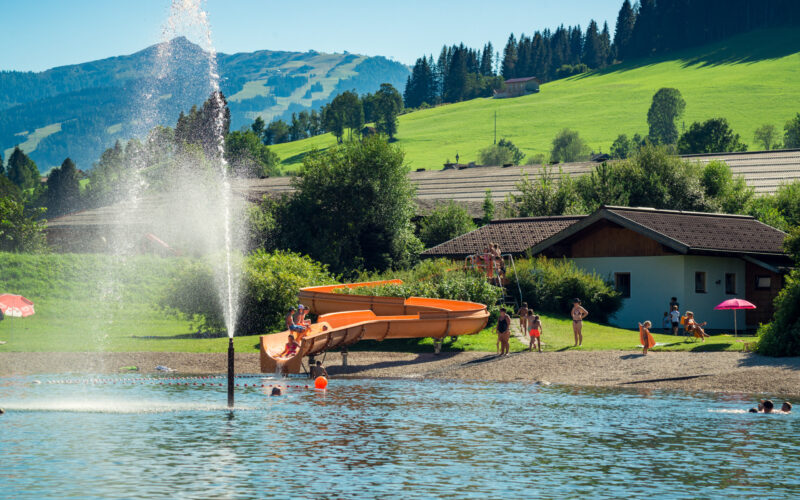 Menschen genießen einen sonnigen Tag am Strand eines Sees mit einer Wasserrutsche, einem Springbrunnen und grasbewachsenen Hügeln im Hintergrund.