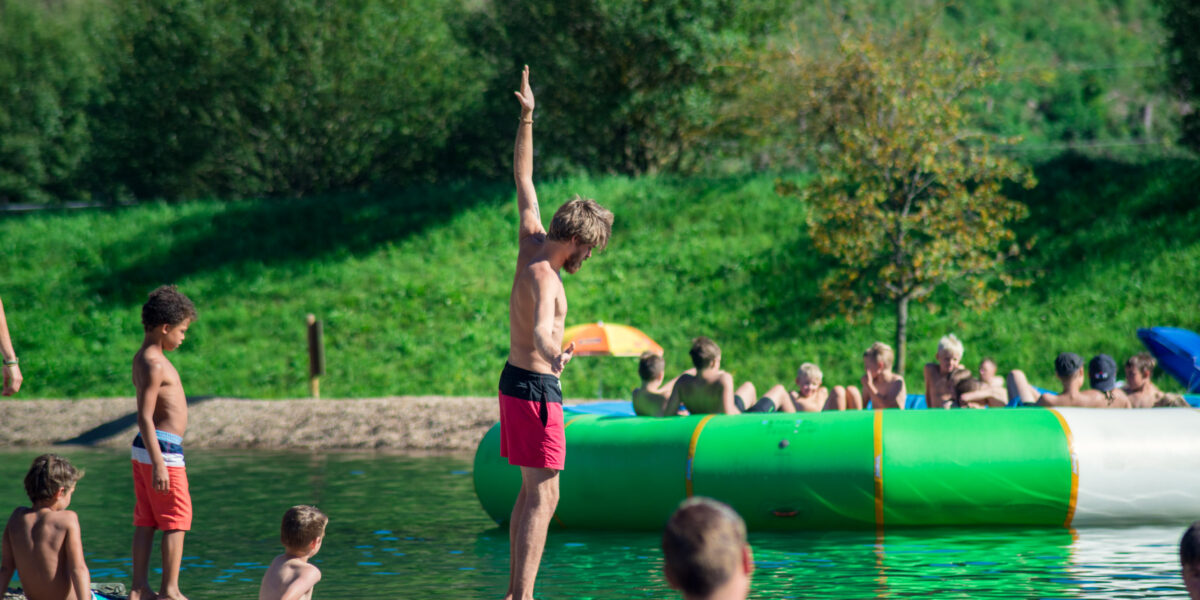 Ein Mann in roter Badehose steht mit erhobenem Arm am Ufer, während Kinder und Erwachsene in der Nähe eines großen aufblasbaren Schwimmkörpers an einem Seeufer spielen.