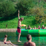 Ein Mann in roter Badehose steht mit erhobenem Arm am Ufer, während Kinder und Erwachsene in der Nähe eines großen aufblasbaren Schwimmkörpers an einem Seeufer spielen.