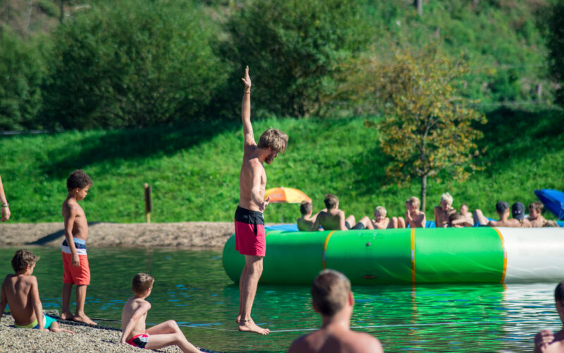 Ein Mann in roter Badehose steht mit erhobenem Arm am Ufer, während Kinder und Erwachsene in der Nähe eines großen aufblasbaren Schwimmkörpers an einem Seeufer spielen.