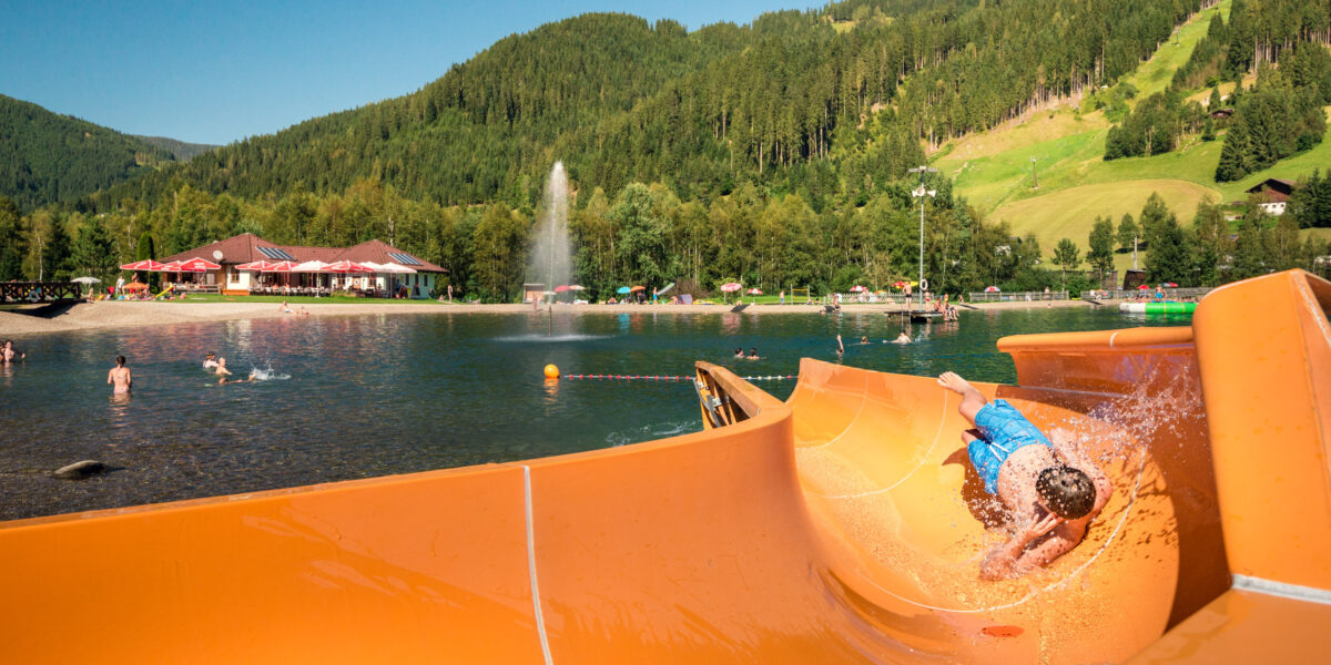Ein Kind rutscht eine orangefarbene Wasserrutsche hinunter in einen natürlichen Badesee, mit schwimmenden Menschen und einer bewaldeten Berglandschaft im Hintergrund.