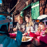Zwei Erwachsene und zwei Kinder in Skikleidung sitzen an einem Holztisch im Freien und genießen das Essen an einem sonnigen Tag neben einem rustikalen Holzgebäude.