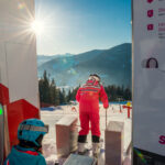 Zwei Kinder in Skiausrüstung bereiten sich an einer Skiliftstation an einem sonnigen Berghang auf eine Abfahrt vor, wobei die Anweisungen auf Schildern in der Nähe stehen.