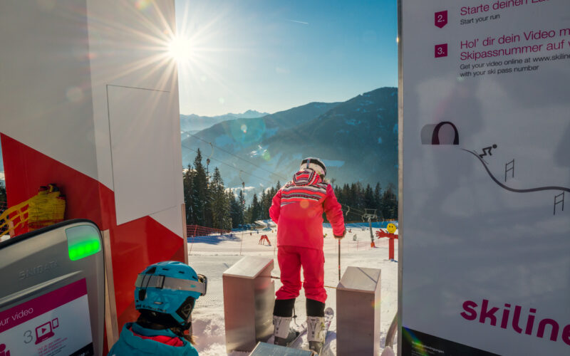 Zwei Kinder in Skiausrüstung bereiten sich an einer Skiliftstation an einem sonnigen Berghang auf eine Abfahrt vor, wobei die Anweisungen auf Schildern in der Nähe stehen.