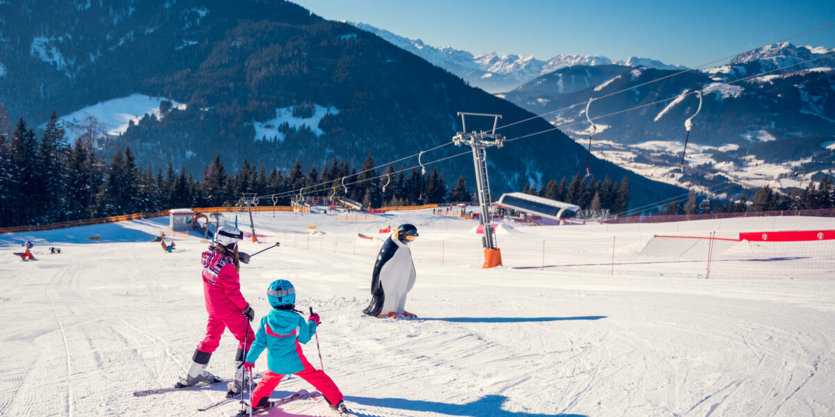 Zwei Kinder in Skiausrüstung üben auf einer verschneiten Piste in der Nähe einer pinguinförmigen Markierung das Skifahren, mit Bergen und Skiliften im Hintergrund.