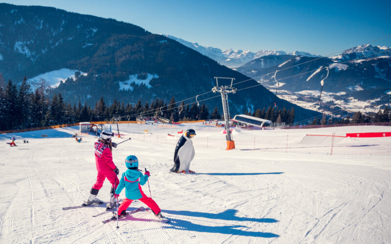 Zwei Kinder in Skiausrüstung üben auf einer verschneiten Piste in der Nähe einer pinguinförmigen Markierung das Skifahren, mit Bergen und Skiliften im Hintergrund.