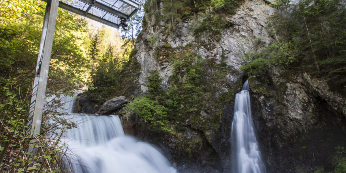 Ein metallener Steg überspannt einen felsigen Wasserfall, der von dichtem grünen Wald und Klippenvegetation umgeben ist.