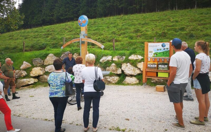 Eine Gruppe von Menschen steht im Freien und hört einem Sprecher in der Nähe eines Informationsdisplays und einer Holzkonstruktion zu, mit einem grasbewachsenen Hügel und Felsen im Hintergrund.