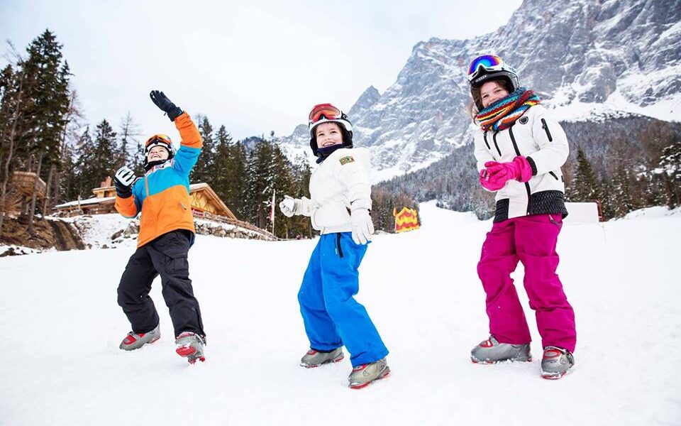 Drei Kinder in Winterkleidung und mit Helmen stehen auf einem verschneiten Hang mit Bergen und Bäumen im Hintergrund.