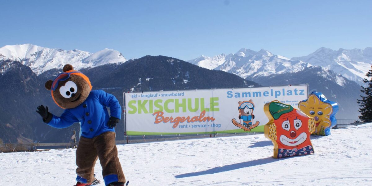 Eine Person in einem Bären-Maskottchen-Kostüm fährt auf einer verschneiten Piste neben einem Schild für eine Skischule Ski, mit Cartoon-Figuren und schneebedeckten Bergen im Hintergrund.
