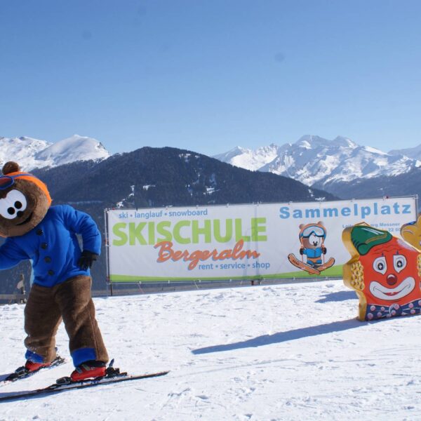 Eine Person in einem Bären-Maskottchen-Kostüm fährt auf einer verschneiten Piste neben einem Schild für eine Skischule Ski, mit Cartoon-Figuren und schneebedeckten Bergen im Hintergrund.