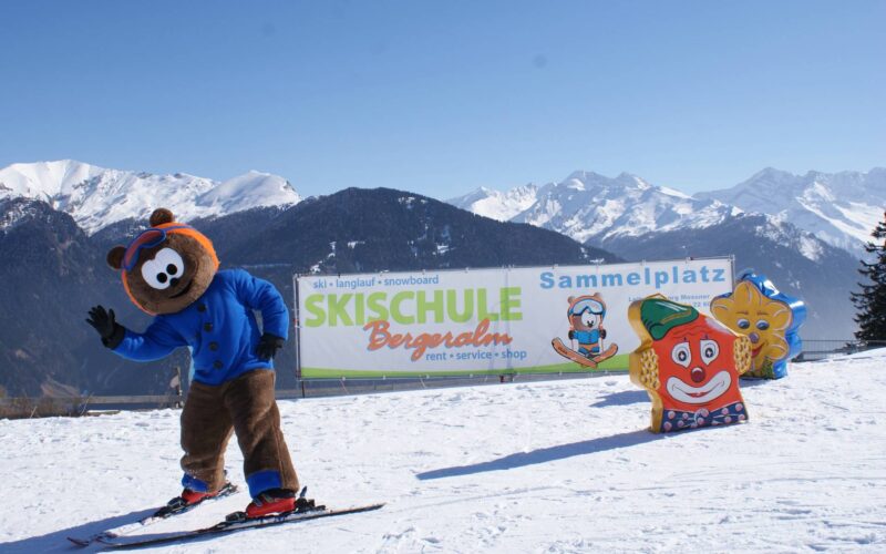 Eine Person in einem Bären-Maskottchen-Kostüm fährt auf einer verschneiten Piste neben einem Schild für eine Skischule Ski, mit Cartoon-Figuren und schneebedeckten Bergen im Hintergrund.