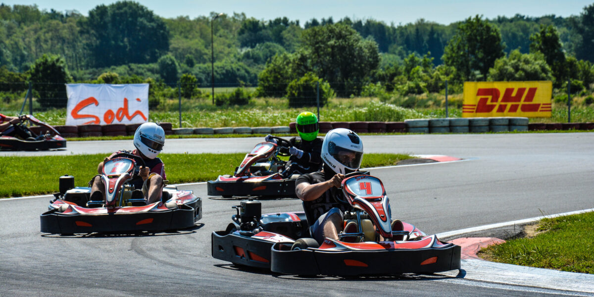 Drei Personen mit Helmen fahren Go-Karts um eine Kurve auf einer Freiluftstrecke, im Hintergrund sind Bäume und Werbebanner zu sehen.