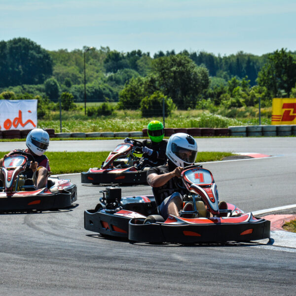 Drei Personen mit Helmen fahren Go-Karts um eine Kurve auf einer Freiluftstrecke, im Hintergrund sind Bäume und Werbebanner zu sehen.