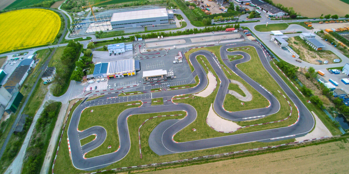 Luftaufnahme einer Go-Kart-Rennstrecke mit gewundenen Asphaltbahnen, umgeben von Gras, Gebäuden, Parkplätzen und Ackerland.