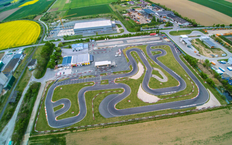 Luftaufnahme einer Go-Kart-Rennstrecke mit gewundenen Asphaltbahnen, umgeben von Gras, Gebäuden, Parkplätzen und Ackerland.