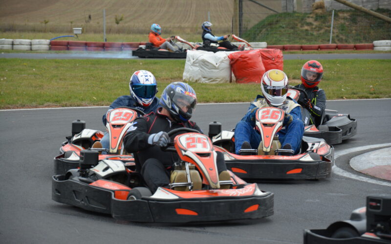 Vier Personen in Rennanzügen und mit Helmen fahren mit Gokarts um eine Kurve, während im Hintergrund zwei weitere Fahrer auf einer Rennstrecke im Freien zu sehen sind.