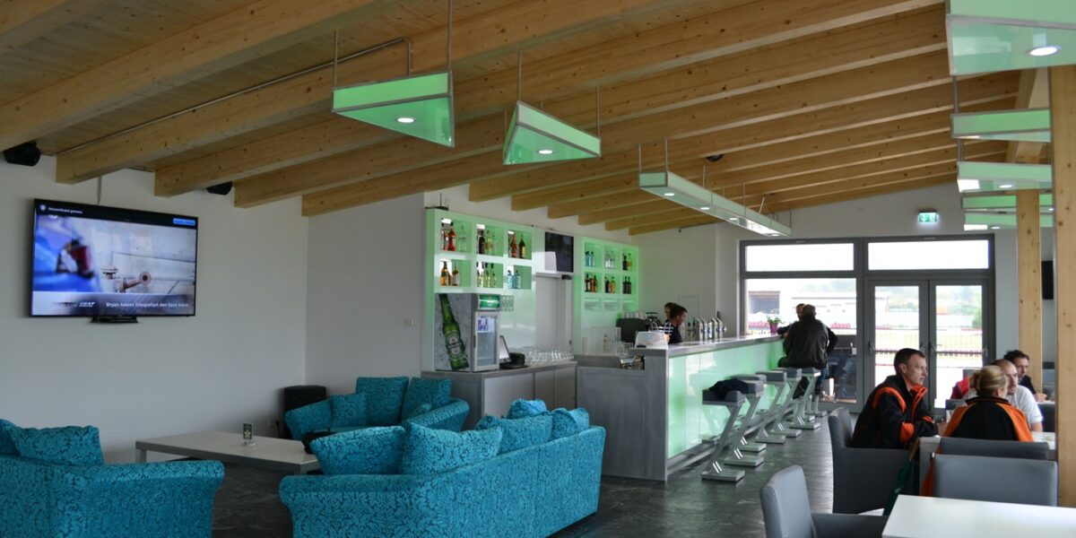 Moderne Lounge mit tealfarbenen Sofas, einem an der Wand montierten Fernseher, einem Barbereich mit Hockern und einigen Tischen unter einer Holzbalkendecke.