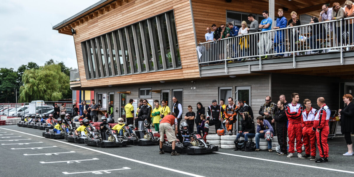 Die Menschen versammeln sich und sehen zu, wie sich die Go-Kart-Fahrer in der Startaufstellung vor einem modernen Holzgebäude mit einem Balkon aufstellen.