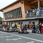 Die Menschen versammeln sich und sehen zu, wie sich die Go-Kart-Fahrer in der Startaufstellung vor einem modernen Holzgebäude mit einem Balkon aufstellen.