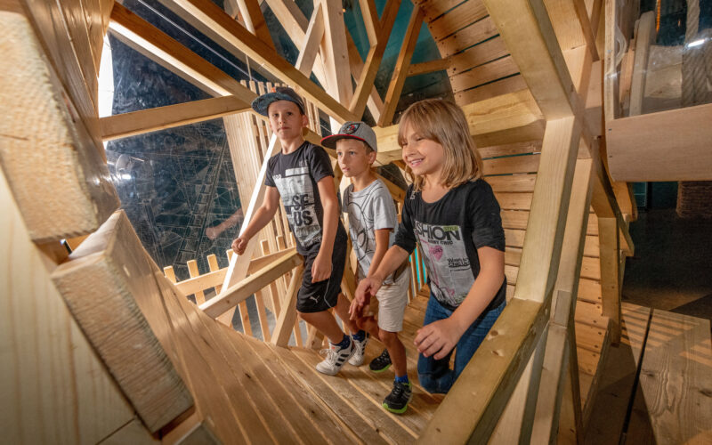 Drei Kinder laufen durch ein hölzernes Spielgerät im Haus, schauen nach vorne und lächeln.