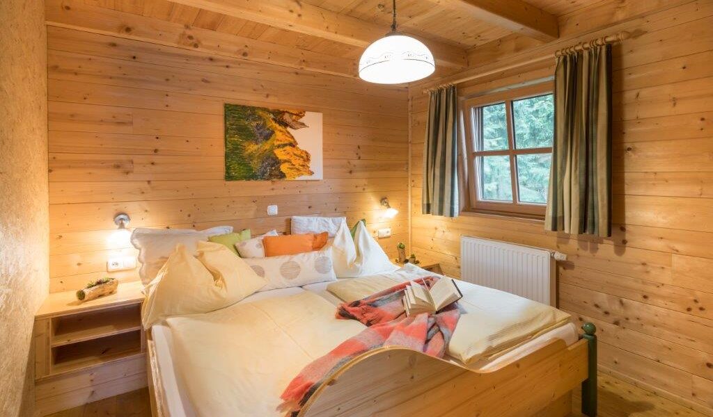 Gemütliches Schlafzimmer aus Holz mit einem Doppelbett, weißer Bettwäsche, Kissen, einem Buch, Nachttischregalen, einem Fenster mit Vorhängen und einem Gemälde an der Wand.
