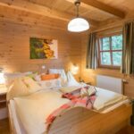 Gemütliches Schlafzimmer aus Holz mit einem Doppelbett, weißer Bettwäsche, Kissen, einem Buch, Nachttischregalen, einem Fenster mit Vorhängen und einem Gemälde an der Wand.