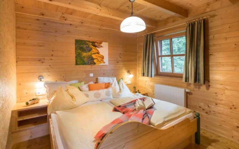 Gemütliches Schlafzimmer aus Holz mit einem Doppelbett, weißer Bettwäsche, Kissen, einem Buch, Nachttischregalen, einem Fenster mit Vorhängen und einem Gemälde an der Wand.